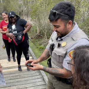 A park ranger holds a small turtle to show it to several teachers standing on a boardwalk. 